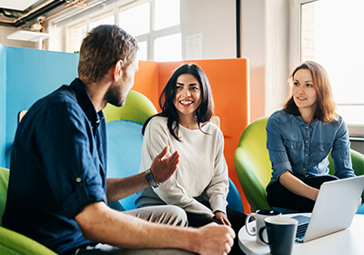 Talent Community.  Three colleagues sitting in colorful lounge chairs, talking and smiling around a table with a laptop and coffee mugs.